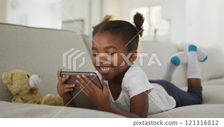 Happy african american girl lying on sofa using digital tablet at home Happy african american girl lying on sofa using digital tablet at home 121316812