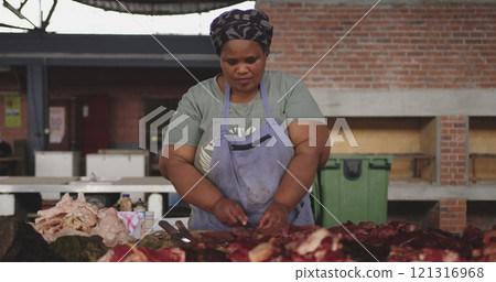 Front view of an African female butcher wearing a headscarf in a township workshop, cutting fresh me 121316968