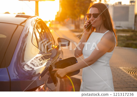 Woman charging electric car while talking on mobile phone during sunset in the street 121317236