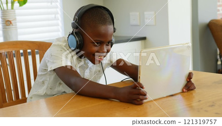 Front view of little black boy playing game on digital tablet at dining table in comfortable home 121319757