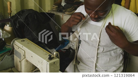 Front view of an African male tailor in a township workshop, sitting by a sewing machine and prepari 121319864