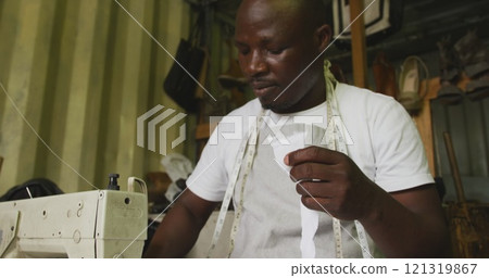 Front view of an African male tailor in a township workshop, sitting by a sewing machine and cutting Front view of an African male tailor in a township workshop, sitting by a sewing machine and cutting 121319867