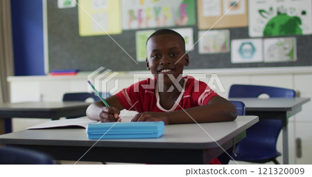 Portrait of happy african american schoolboy sitting at classroom, making notes, looking at camera Portrait of happy african american schoolboy sitting at classroom, making notes, looking at camera 121320009