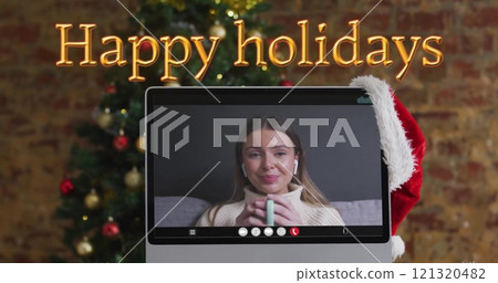 Image of happy holidays text over christmas tree and caucasian woman on laptop screen 121320482