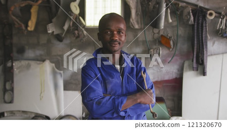 Portrait of a happy African male panel beater in a township workshop, holding a wrench, looking at c 121320670