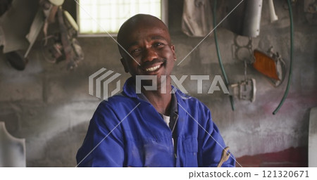 Portrait of a happy African male panel beater in a township workshop, holding a wrench, looking at c 121320671