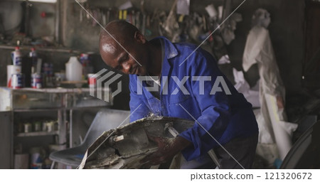 Front view of an African male panel beater in a township workshop, sanding and preparing surface of 121320672
