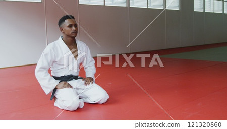 SIde view of a teenage biracial male judoka wearing white judogi, kneeling on mats in the gym before SIde view of a teenage biracial male judoka wearing white judogi, kneeling on mats in the gym before 121320860