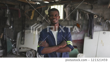 Portrait of a happy African male panel beater in a township workshop, looking at camera with arms cr Portrait of a happy African male panel beater in a township workshop, looking at camera with arms cr 121321459