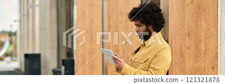 African American man standing outdoors, using tablet while leaning against wooden wall, appearing focused and engaged 121321878