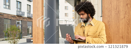 Man standing outdoors working on laptop while leaning against wooden wall in urban setting showing concentration. Man is absorbed in work 121321879