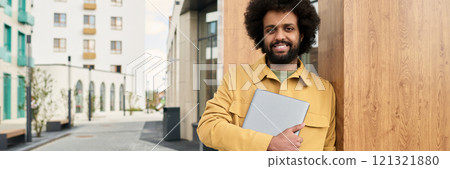 Smiling man with Afro hairstyle holding laptop while standing outdoor in urban environment. Displaying confident posture and casual attire Smiling man with Afro hairstyle holding laptop while standing outdoor in urban environment. Displaying confident posture and casual attire 121321880