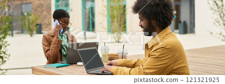 Colleagues working at outdoor table with laptops and beverages, engaging in conversation. One colleague multitasking while using a phone and wearing glasses 121321925