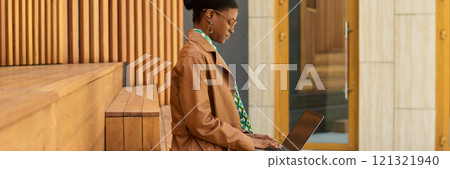Woman wearing a brown jacket and colorful scarf, working on a laptop while sitting in a modern, well-designed outdoor space with wooden and architectural elements around her 121321940