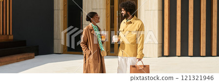 Two professionals standing outside modern building entrance having conversation while holding disposable cups, one holding a briefcase, wearing business attire showing engagement 121321946