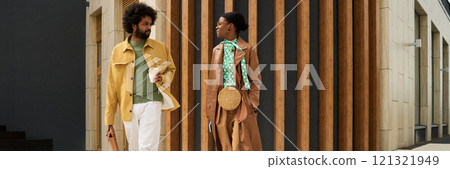 Two people engaged in conversation while holding beverages and walking along a modern building with wooden paneling Two people engaged in conversation while holding beverages and walking along a modern building with wooden paneling 121321949
