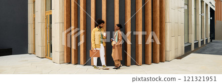 Two business professionals engaging in conversation outside modern office building while holding coffee cups and briefcase. Wooden panel backdrop accents corporate environment Two business professionals engaging in conversation outside modern office building while holding coffee cups and briefcase. Wooden panel backdrop accents corporate environment 121321951