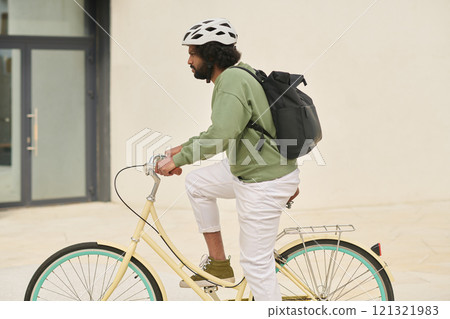 Man riding bicycle on sunny urban street carrying black backpack on shoulders wearing white safety helmet ensuring safety. Door of modern building visible in background Man riding bicycle on sunny urban street carrying black backpack on shoulders wearing white safety helmet ensuring safety. Door of modern building visible in background 121321983
