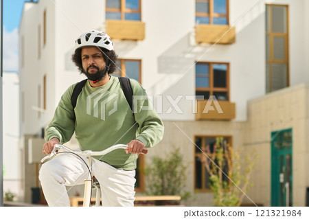 Person riding bicycle in urban area wearing helmet and green sweater. Residential buildings with balconies in background enhance the cityscape look 121321984