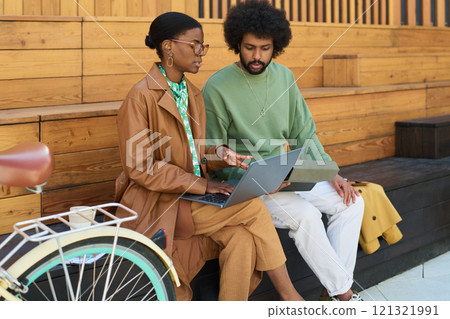 Two colleagues collaborating on laptop while sitting on wooden bench. Woman wearing glasses and coat discussing with her teammate who is wearing green sweatshirt Two colleagues collaborating on laptop while sitting on wooden bench. Woman wearing glasses and coat discussing with her teammate who is wearing green sweatshirt 121321991