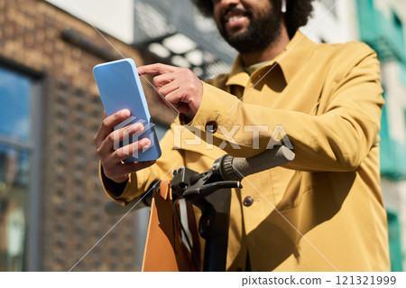 Man holding smartphone while standing on street and smiling with scooter beside him. Cloudless sky reflecting on window of nearby building 121321999