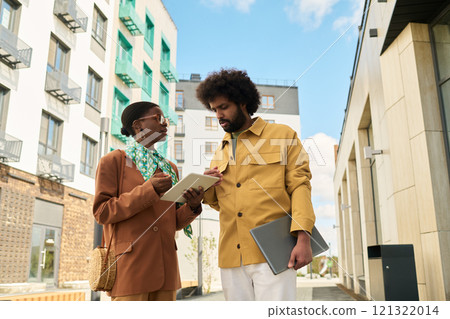 Two professionals discussing business on street with modern buildings in background. Urban setting enhances contemporary vibe, while tablet and notebook signify serious work Two professionals discussing business on street with modern buildings in background. Urban setting enhances contemporary vibe, while tablet and notebook signify serious work 121322014