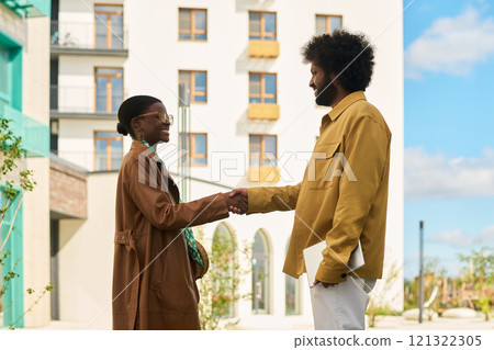 Two individuals engaging in handshake in outdoor urban setting with high-rise buildings in background. Both are smiling, conveying mutual respect and friendliness 121322305