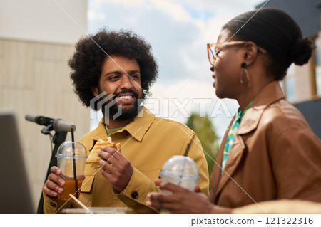 Man with curly hair wearing a yellow jacket enjoying a drink and snack while engaging in conversation with a woman in glasses, both smiling and relaxed 121322316