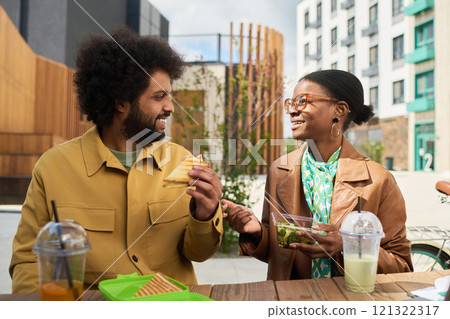 Two friends sitting at an outdoor cafe enjoying lunch together. They are engaged in conversation and sharing food, creating an atmosphere of camaraderie and relaxation 121322317