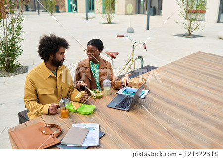 Colleagues conversing while sitting at outdoor table, reviewing documents. Brown leather bag and bicycle in background, suggesting casual yet professional setting 121322318