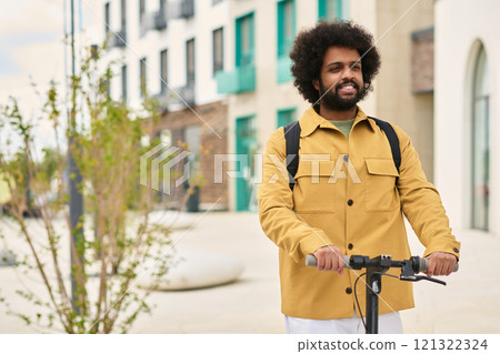 Man smiling and holding bicycle in urban setting with modern apartment buildings in background wearing brown jacket and white pants 121322324