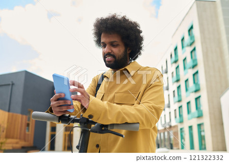 African American man standing outdoors using smartphone while holding handlebar of scooter captured in urban environment with modern buildings in background 121322332