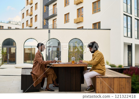 Two individuals having work meeting at outdoor workspace in front of modern building complex. Engaging in productive conversation while seated at wooden table 121322418