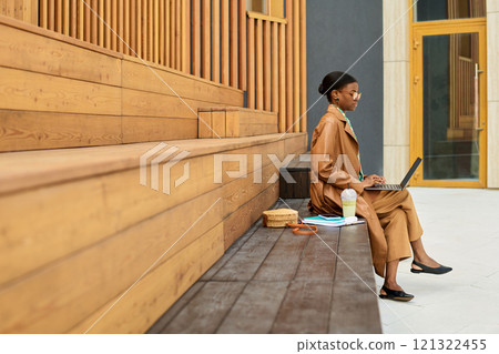 Woman sitting on wooden bench using laptop and drinking from coffee cup in an outdoor urban setting. Sunlight shining and creating shadows on wooden panels of the building behind her Woman sitting on wooden bench using laptop and drinking from coffee cup in an outdoor urban setting. Sunlight shining and creating shadows on wooden panels of the building behind her 121322455