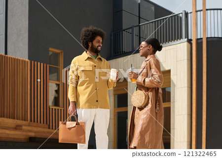 Young man and woman standing outdoors in front of a modern building, engaging in conversation with coffee cups in hands, both dressed stylishly with casual elegance 121322465