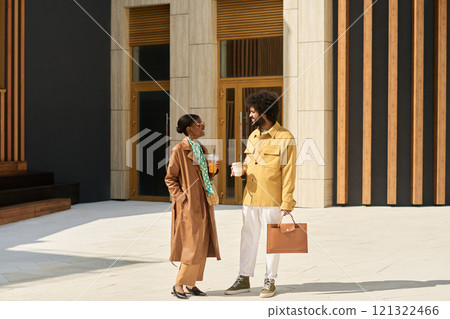 African American man and Black woman engaging in conversation while walking under sunny weather holding coffee cups and briefcase at modern office building 121322466