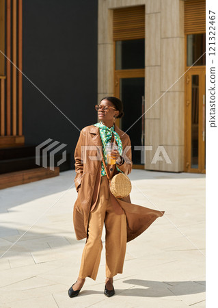 Person showcasing stylish outfit while walking outdoors in urban setting. Featuring long coat, sunglasses, and carrying woven bag in hand while integrating green scarf 121322467