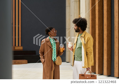 Two individuals are engaging in conversation, each holding coffee cups, in an indoor setting with modern architectural elements. Both are wearing stylish coats and accessories, indicating a 121322468