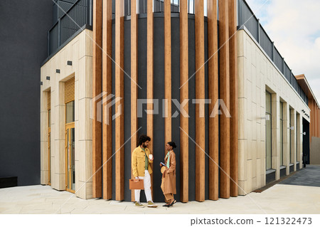 Two people having conversation outside contemporary structure with wooden accents and tall vertical elements. Modern design and clean lines define the architecture Two people having conversation outside contemporary structure with wooden accents and tall vertical elements. Modern design and clean lines define the architecture 121322473