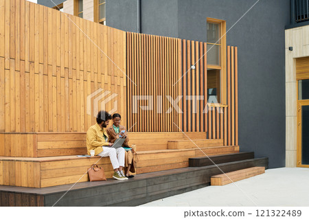 Two business people sitting on wooden steps outside modern building, talking and relaxing, background consists of contemporary architecture Two business people sitting on wooden steps outside modern building, talking and relaxing, background consists of contemporary architecture 121322489