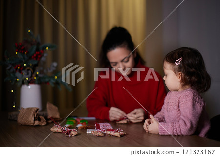 mother and her little child girl together wrapping Christmas gifts at home in the evening, preparing presents for family. 121323167