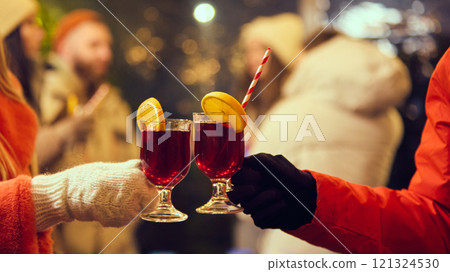 Two glasses of mulled wine with orange slices and striped straws clinking together, celebrating a cozy winter evening under festive lights. 121324530