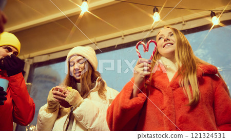Young cheerful people in warm winter clothes, gathering outdoors to celebrate winter holidays, attending fairs. Girls with candies and hot drinks 121324531