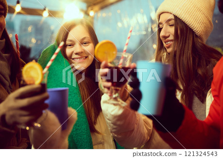 Young smiling women wearing warm hats and scarves drinking hot drinks, mulled wine. Friends gathering at outdoor winter fair Young smiling women wearing warm hats and scarves drinking hot drinks, mulled wine. Friends gathering at outdoor winter fair 121324543