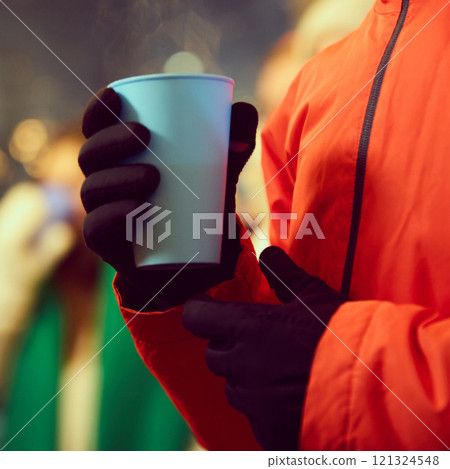 Close-up view of hand wearing black gloves holding blue cup with hot drink, set against blurred festive background. Outdoor fairs during holidays 121324548
