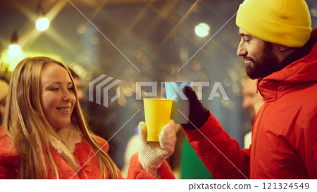 Bearded man in yellow beanie and smiling woman in orange coat sharing warm moment toasting with colorful cups during festive outdoor winter celebration. Bearded man in yellow beanie and smiling woman in orange coat sharing warm moment toasting with colorful cups during festive outdoor winter celebration. 121324549