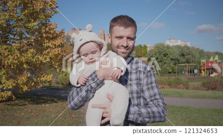 A joyful father lovingly holds his adorable baby, who is dressed in a cozy outfit, in a beautiful sunny park setting 121324653