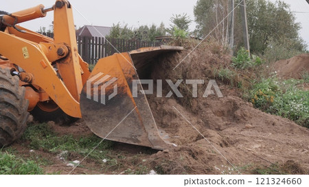 An excavator bucket is actively lifting dirt at a construction site which is surrounded by lush greenery 121324660