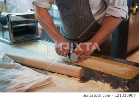 A chef prepares pasta by cutting and rolling the raw dough on a metal countertop. A chef prepares pasta by cutting and rolling the raw dough on a metal countertop. 121326907