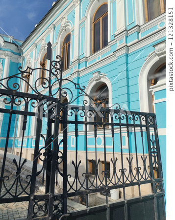 A close-up view of an intricately designed wrought-iron gate framing the blue facade of a historic building with decorative arches and large windows 121328151